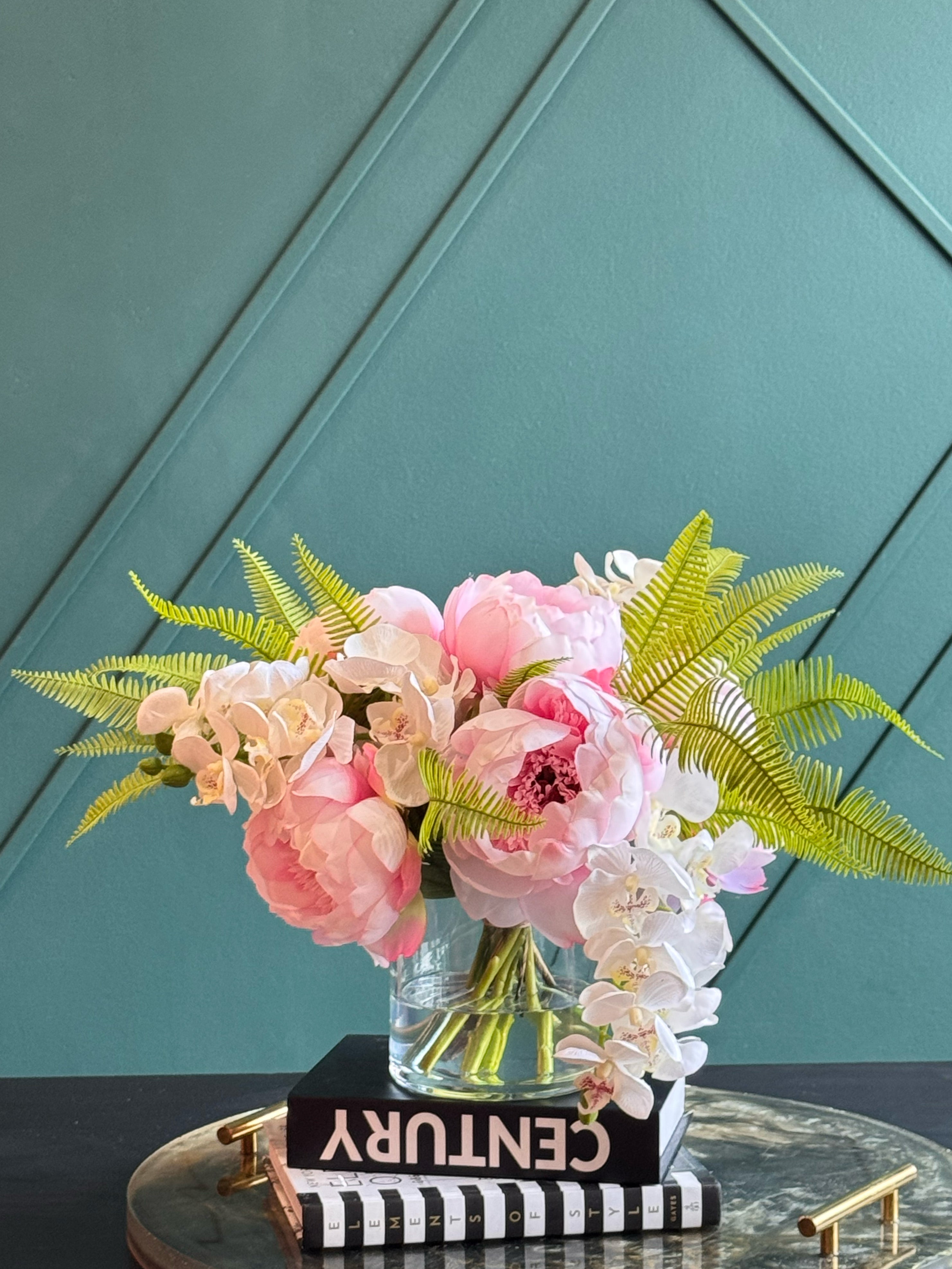 Handcrafted Real Touch pink peony and white orchid arrangement displayed in a clear glass vase with lush green fern foliage.