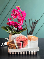 A styled gift set on a white tray featuring a vibrant pink artificial orchid, a reed diffuser with dark sticks, a round container of pink bath salts