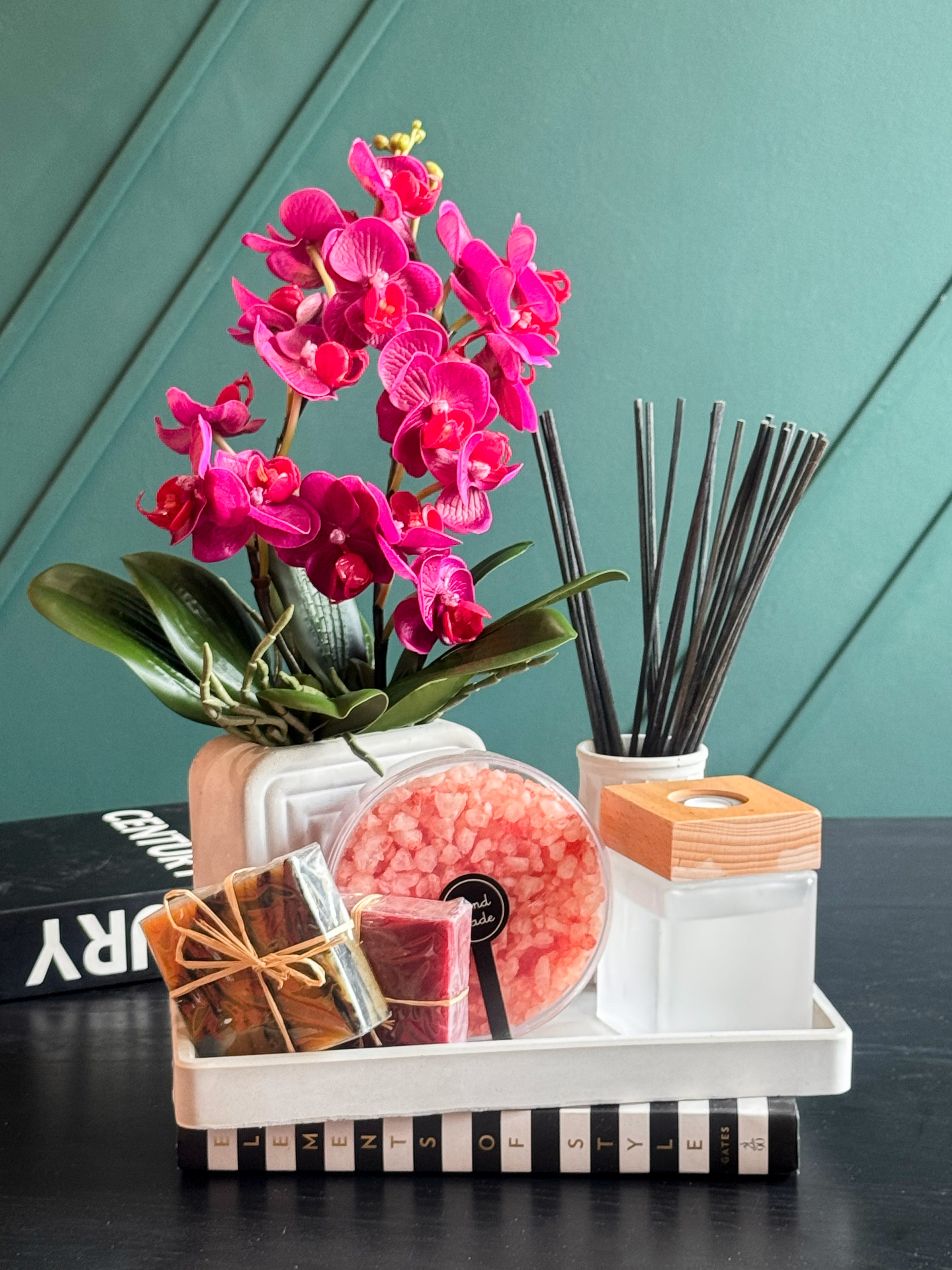 A styled gift set on a white tray featuring a vibrant pink artificial orchid, a reed diffuser with dark sticks, a round container of pink bath salts