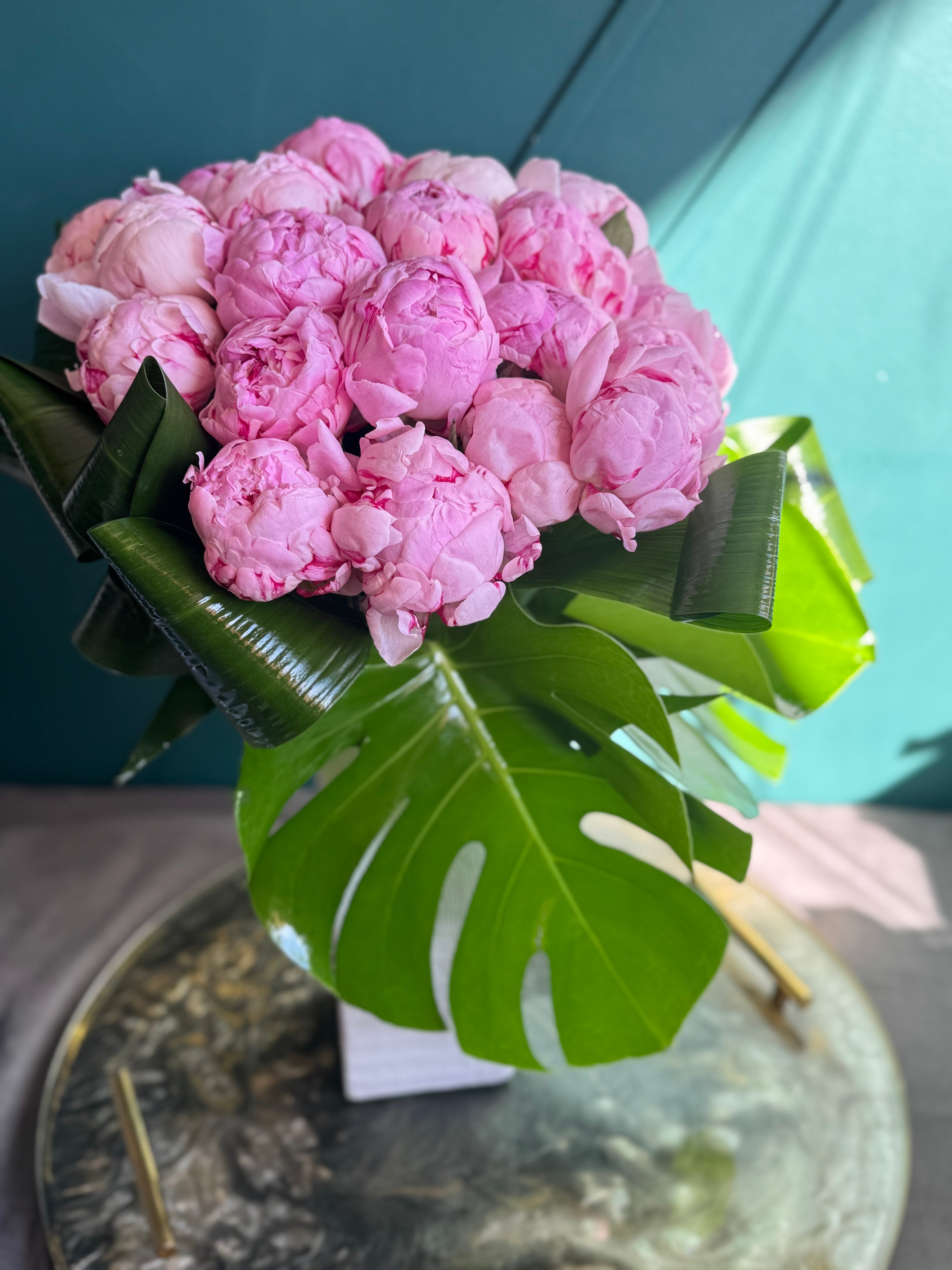 Two dozen pink peonies with monstera leaves in modern square vase